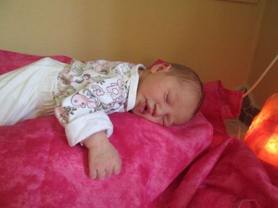 Ein Baby schläft auf einer rosa Decke mit einer Lampe in der Nähe. Das Baby trägt ein weißes Strampler mit Blumendesign.