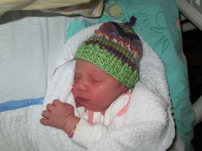 Ein Neugeborenes schläft friedlich in einem Krankenhausbett und trägt eine bunte gestrickte Mütze. Das Baby ist in eine weiße Decke gewickelt.