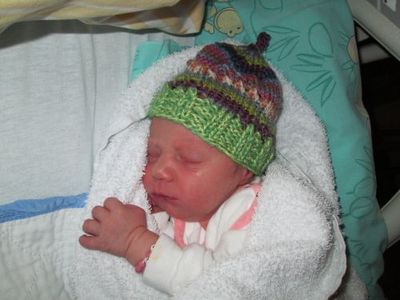 Ein Neugeborenes schläft friedlich in einem Krankenhausbett und trägt eine bunte gestrickte Mütze. Das Baby ist in eine weiße Decke gewickelt.