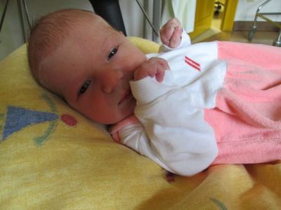 Ein Babymädchen liegt auf einer gelben Decke mit einem rosa Outfit und schaut neugierig in die Kamera.