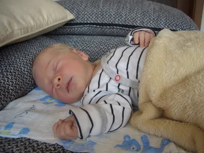 Ein Baby schläft friedlich auf einem Bett, bedeckt mit einer blau-weißen Decke mit Bären-Cartoons. Das Baby trägt ein gestreiftes Outfit und ruht auf einer beigen Decke.