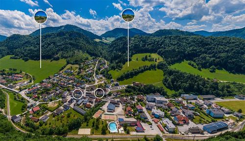 Luftaufnahme eines Dorfes zwischen Bergen. Das Dorf hat mehrere Gebäude, darunter Häuser, eine Schule und ein Schwimmbad. Das Gebiet ist von üppigen grünen Feldern und Bäumen umgeben. Der Himmel ist blau mit einigen Wolken.