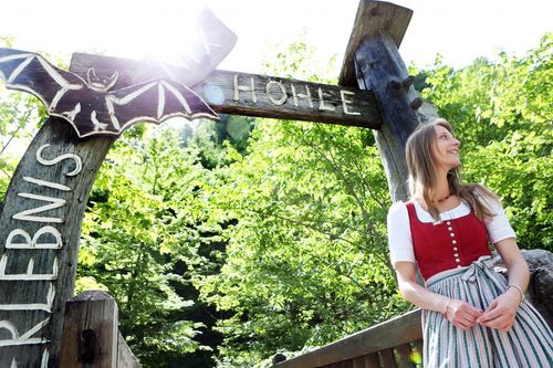 Eine lächelnde Frau in traditioneller deutscher Kleidung steht neben einem hölzernen Schild mit einer Fledermaus-Design, das 'Hohle' liest. Sie ist von üppigen grünen Bäumen umgeben.