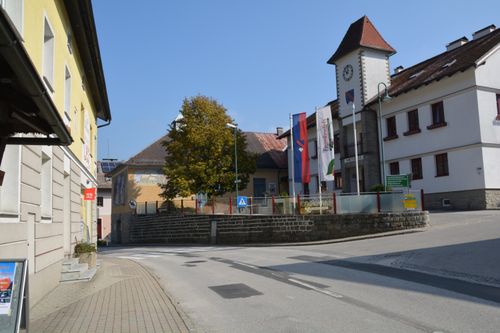 Ein Straßenblick mit Turm, Baum und Flaggen. Gebäude säumen die Straße mit rotem Dach. Eine Uhr ist auf dem Turm sichtbar.