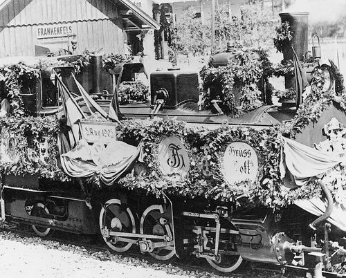 Eine alte Dampflokomotive mit Blumen und Bannern steht auf den Gleisen neben einem Gebäude. Das Schild sagt FRANKENFELS.