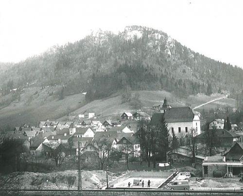 Ein Schwarz-Weiß-Foto einer Stadt mit einem Berg im Hintergrund, einer Kirche und Häusern. Es gibt Menschen in der Nähe einer Baustelle.