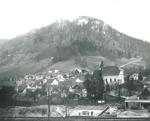 Ein Schwarz-Weiß-Foto einer Stadt mit einem Berg im Hintergrund, einer Kirche und Häusern. Es gibt Menschen in der Nähe einer Baustelle.