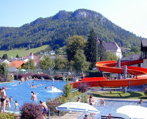 Ein lebhaftes Schwimmbad mit Schwimmern und anderen, die Wasserrutschen genießen, vor einer Kulisse aus üppigem Grün und Bergen.