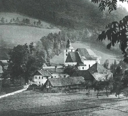 Ein historisches Schwarz-Weiß-Foto zeigt ein Dorf mit einer markanten Kirche, umgeben von Bäumen, Feldern und Hügeln.