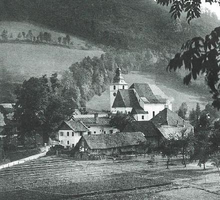 Ein historisches Schwarz-Weiß-Foto zeigt ein Dorf mit einer markanten Kirche, umgeben von Bäumen, Feldern und Hügeln.