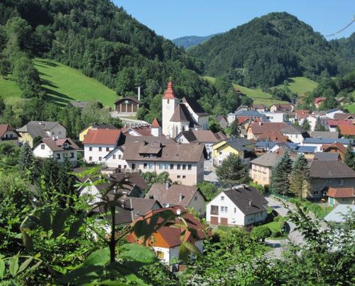 Ein in den Bergen gelegenes Dorf mit einer Kirche im Zentrum, umgeben von üppiger Grünfläche und Häusern in verschiedenen Farben.