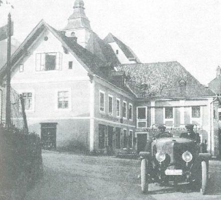 Ein altes Foto zeigt ein Oldtimer-Auto mit zwei Männern, die es vor einem großen Gebäude mit vielen Fenstern und einem Turm fahren.