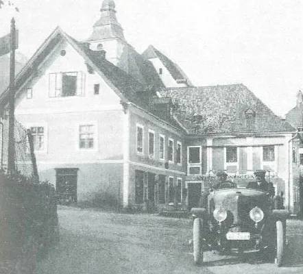Ein altes Foto zeigt ein Oldtimer-Auto mit zwei Männern, die es vor einem großen Gebäude mit vielen Fenstern und einem Turm fahren.