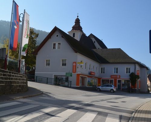 Ein weißes Gebäude mit orangefarbenen Akzenten, einem Turm und einer Uhr steht neben einer Straße, mit einem silbernen Auto davor geparkt.