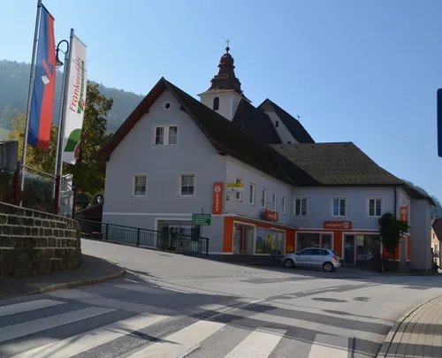 Ein weißes Gebäude mit orangefarbenen Akzenten, einem Turm und einer Uhr steht neben einer Straße, mit einem silbernen Auto davor geparkt.