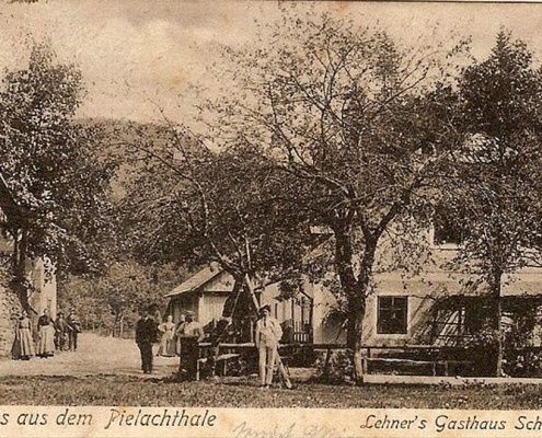 Ein altes Foto zeigt mehrere Personen vor einem Haus mit Bäumen. Das Haus trägt den Namen 'Lehners Gasthaus'.
