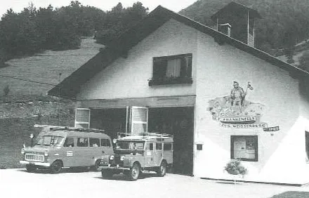 Ein Schwarz-Weiß-Foto einer Garage mit Logo. Zwei Fahrzeuge sind davor geparkt. Ein Van und ein Jeep mit Gepäck auf dem Dach.