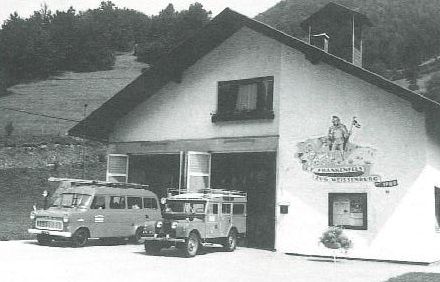 Ein Schwarz-Weiß-Foto einer Garage mit Logo. Zwei Fahrzeuge sind davor geparkt. Ein Van und ein Jeep mit Gepäck auf dem Dach.