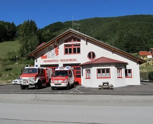 Ein Feuerwehrhaus mit rotem Dach und Fenstern steht vor einem Berg. Zwei Feuerwehrfahrzeuge sind davor geparkt.