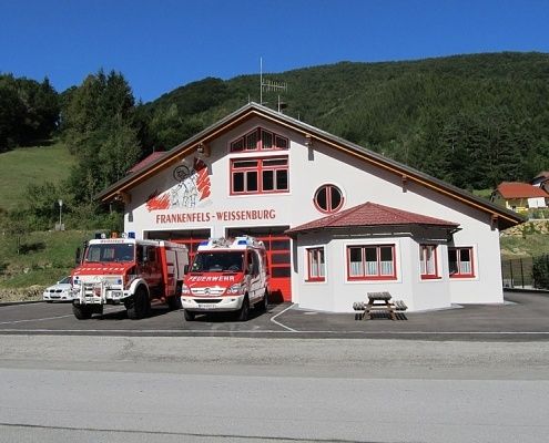 Ein Feuerwehrhaus mit rotem Dach und Fenstern steht vor einem Berg. Zwei Feuerwehrfahrzeuge sind davor geparkt.