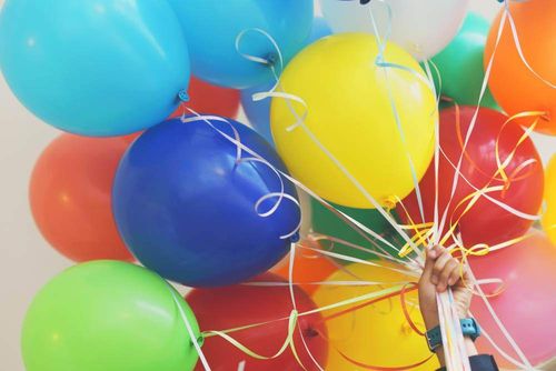 Eine Person hält eine Gruppe bunter Luftballons, die jeweils mit Bändern befestigt sind. Die Ballons sind in verschiedenen Farben, darunter blau, gelb, rot und grün.
