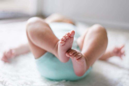 Ein Baby liegt auf einer weißen Oberfläche, trägt eine Windel und hat beide Beine angehoben, wobei kleine Füße zu sehen sind.