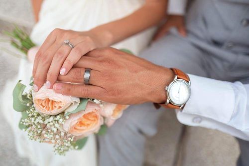 Eine Braut und ein Bräutigam halten während ihrer Hochzeitszeremonie Händchen. Die Braut hält einen Strauß Rosen und Babys Atmen, während der Bräutigam eine Uhr trägt.