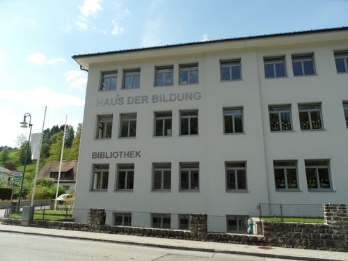 Ein großes weißes Gebäude mit der Aufschrift Haus der Bildung Bibliothek, vielen Fenstern und einem geparkten Auto davor.
