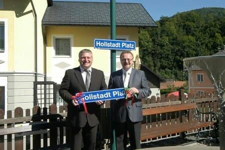 Zwei Männer in Anzügen halten ein Schild mit der Aufschrift 'Hollstaedt Platz' vor einem gelben Haus. Dahinter sind ein Straßenschild und ein Zaun zu sehen.