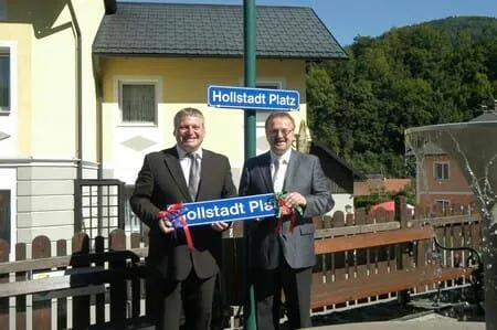 Zwei Männer in Anzügen halten ein Schild mit der Aufschrift 'Hollstaedt Platz' vor einem gelben Haus. Dahinter sind ein Straßenschild und ein Zaun zu sehen.