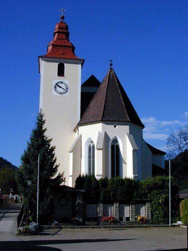 Eine weiße Kirche mit einer Uhr auf ihrem Turm ist von Büschen und Bäumen umgeben unter einem blauen Himmel.