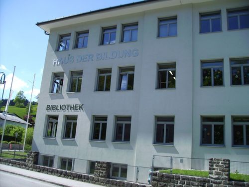 Ein weißes Gebäude mit den Worten 'Haus der Bildung' und 'Bibliothek' darauf. Das Gebäude hat mehrere Fenster und einen Zaun.