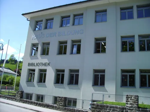 Ein weißes Gebäude mit den Worten 'Haus der Bildung' und 'Bibliothek' darauf. Das Gebäude hat mehrere Fenster und einen Zaun.