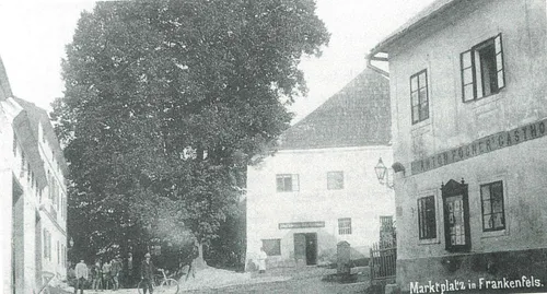 Ein altes Foto zeigt einen Mann, der in der Nähe eines Gebäudes mit mehreren Fenstern und einem Tor ein Fahrrad fährt. Eine Frau steht in der Nähe des Gebäudes.