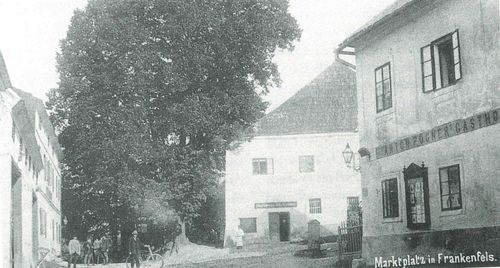 Ein altes Foto zeigt einen Mann, der in der Nähe eines Gebäudes mit mehreren Fenstern und einem Tor ein Fahrrad fährt. Eine Frau steht in der Nähe des Gebäudes.