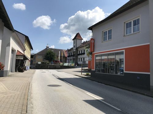 Eine ruhige Straße mit Gebäuden und einem Glockenturm. Draußen sind Fahnen und Pflanzen. Der Himmel ist bewölkt.