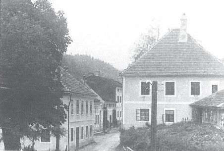 Ein Schwarz-Weiß-Bild einer Dorfstraße mit Häusern und einem Pfad. Bäume säumen die Seiten, und auf einem Haus ist ein Schornstein zu sehen.