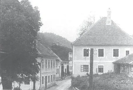 Ein Schwarz-Weiß-Bild einer Dorfstraße mit Häusern und einem Pfad. Bäume säumen die Seiten, und auf einem Haus ist ein Schornstein zu sehen.