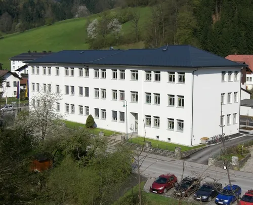 Eine Vogelperspektive eines großen weißen Gebäudes mit mehreren Fenstern, vor dem Hintergrund grüner Hügel und Bäume. Vor dem Gebäude sind mehrere Autos geparkt.