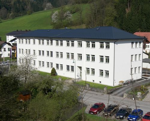 Eine Vogelperspektive eines großen weißen Gebäudes mit mehreren Fenstern, vor dem Hintergrund grüner Hügel und Bäume. Vor dem Gebäude sind mehrere Autos geparkt.