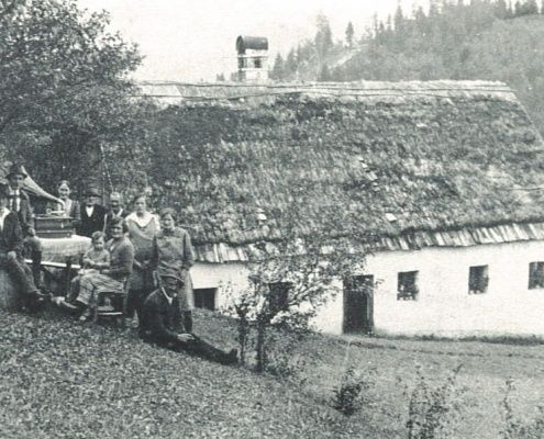 Schwarz-Weiß-Foto einer Gruppe von Menschen, die vor einem Haus mit Strohdach und Schornstein stehen und sitzen.