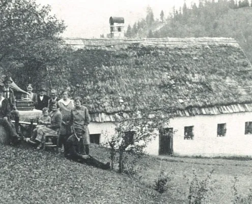 Schwarz-Weiß-Foto einer Gruppe von Menschen, die vor einem Haus mit Strohdach und Schornstein stehen und sitzen.