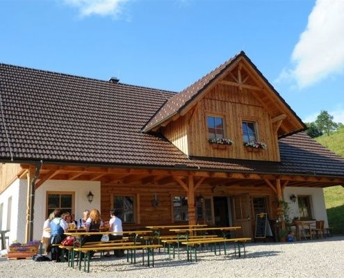 Eine Gruppe von Menschen sitzt an Tischen vor einem Holzhaus mit braunem Dach. Das Haus hat Glasfenster und ist von Pflanzen und Bäumen umgeben.