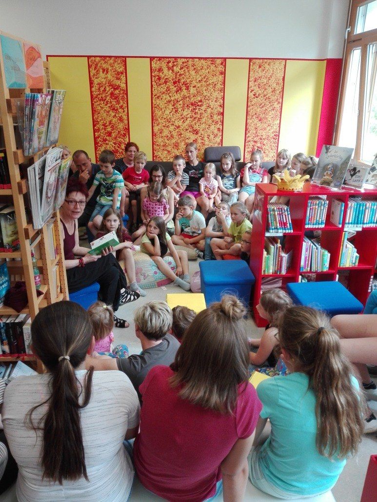 In einer Bibliothek sitzt eine Gruppe von Kindern um eine Frau, die ein Buch liest. Bunte Wanddekorationen und Bücherregale mit Büchern sind vorhanden.