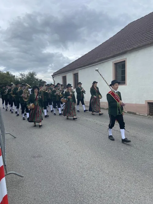 Ein Marschkapelle in traditionellen grünen und weißen Uniformen spielt Instrumente, darunter Tubas, Trompeten und Flöten, während sie eine Straße entlang laufen.