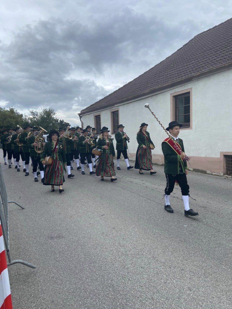 Ein Marschkapelle in traditionellen grünen und weißen Uniformen spielt Instrumente, darunter Tubas, Trompeten und Flöten, während sie eine Straße entlang laufen.
