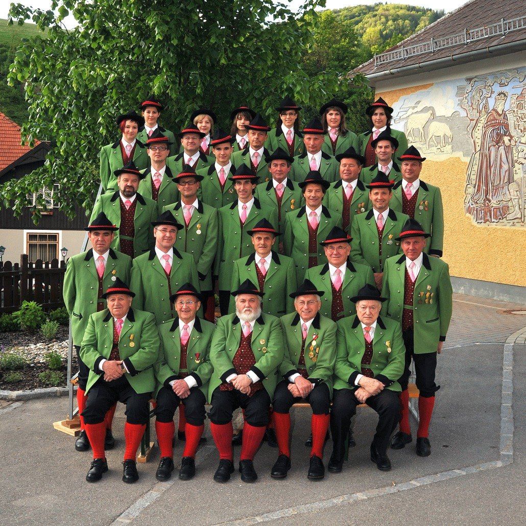 Eine Gruppe von Männern, gekleidet in traditionelle grüne und schwarze Uniformen mit roten Socken, sitzen und stehen vor einem Gebäude mit einer Wandmalerei.