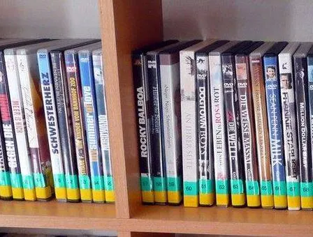 Ein hölzerne Regalplatte zeigt eine Reihe von DVDs mit gelben und grünen Bändern am unteren Rand, die verschiedene Filmtitel anzeigen.