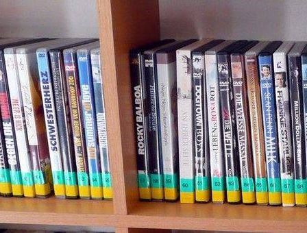 Ein hölzerne Regalplatte zeigt eine Reihe von DVDs mit gelben und grünen Bändern am unteren Rand, die verschiedene Filmtitel anzeigen.