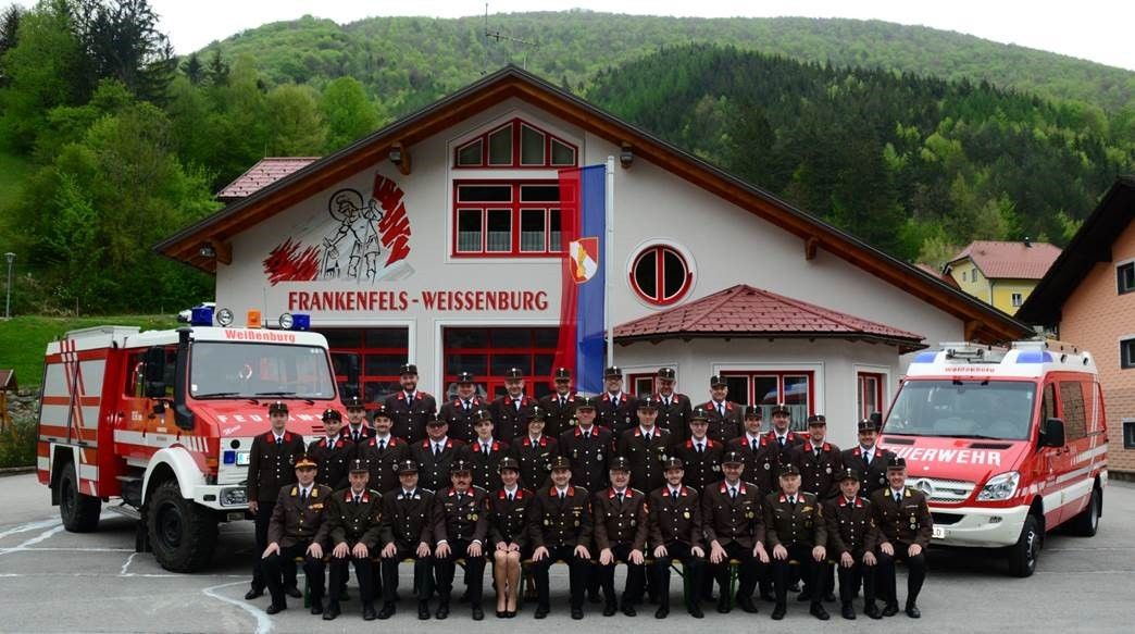 Eine Gruppe von Feuerwehrleuten posiert für ein Foto vor einem Gebäude mit Flaggen und Feuerwehrfahrzeugen.